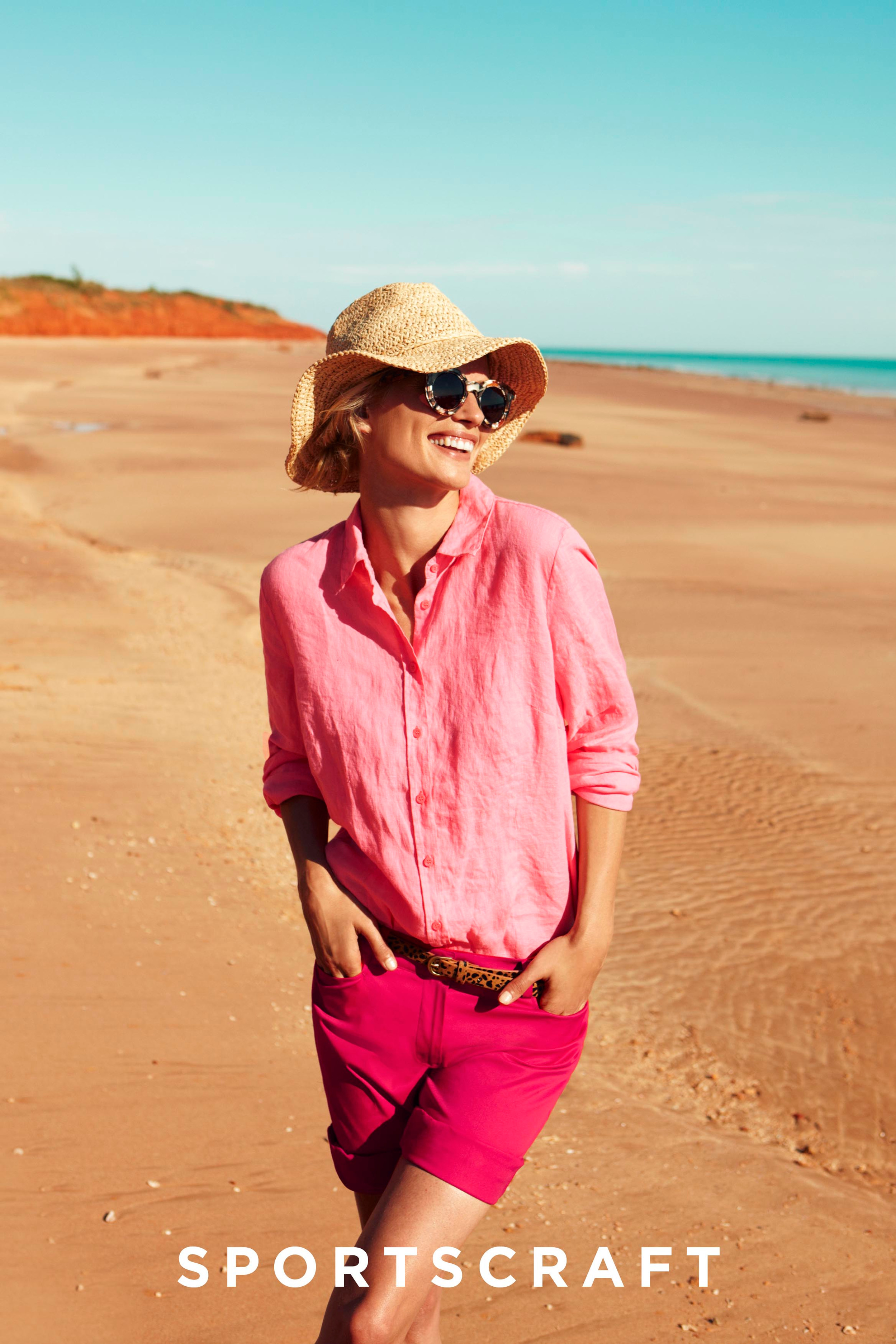 A happy woman in a pink linen shirt and shorts poses on a sandy beach with a straw hat and sunglasses on a sunny day.