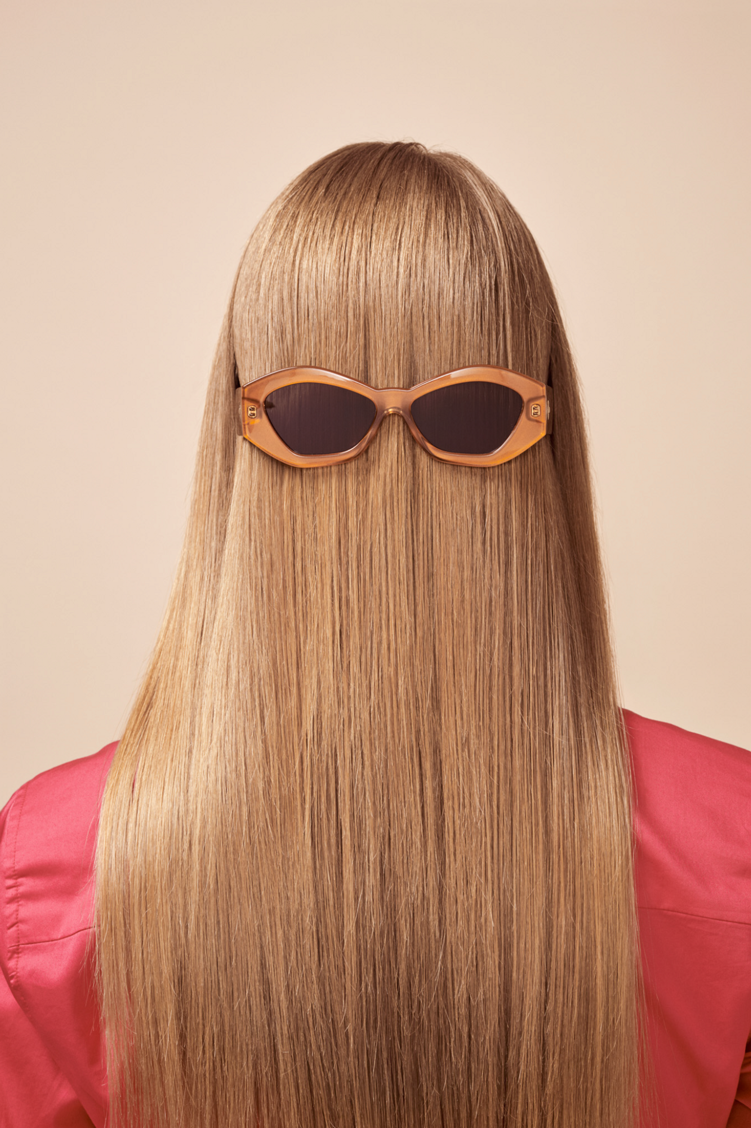 Back view of a woman with long blonde hair wearing peach sunglasses. Focus is on hairstyle and eyewear against a peach background.
