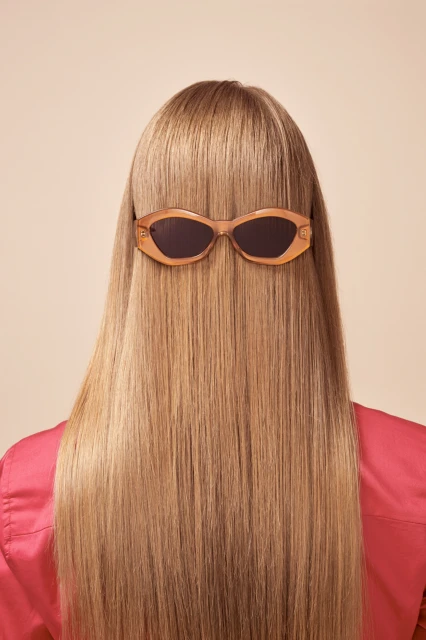 Back view of a woman with long blonde hair wearing peach sunglasses. Focus is on hairstyle and eyewear against a peach background.