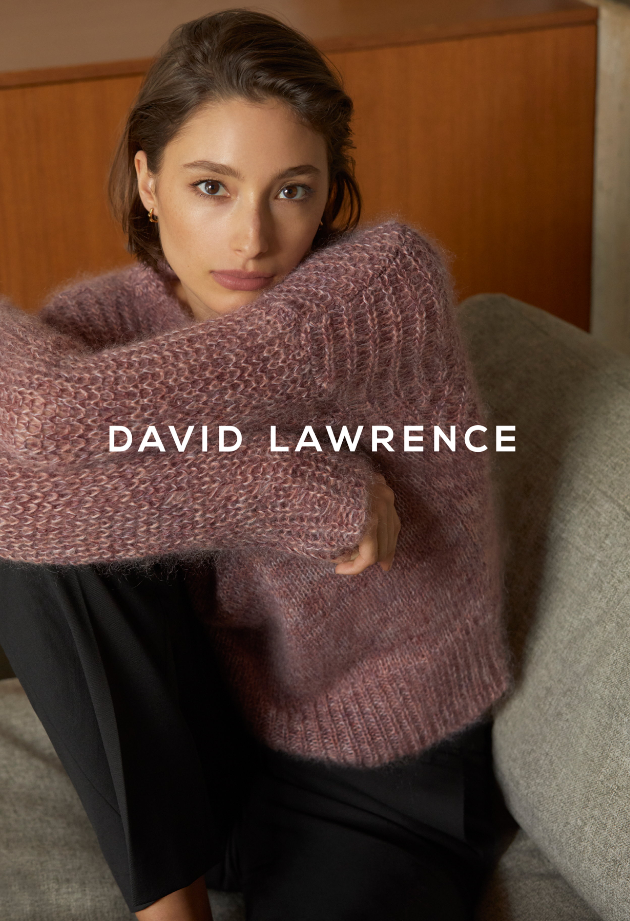 Woman in pink sweater poses indoors on gray couch. Fashion shot with soft lighting, cozy textures, and the brand name DAVID LAWRENCE.