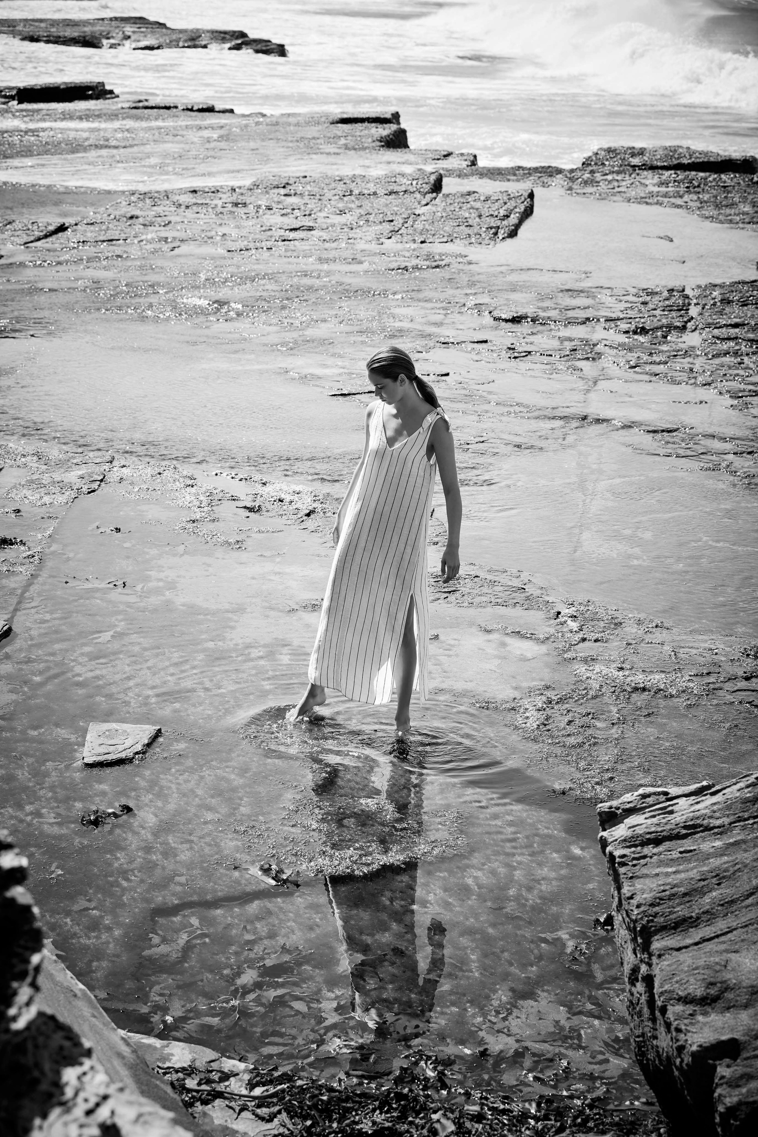 Black and white shot of a woman in a striped dress wading in shallow water with a rocky shoreline and ocean waves in the background.