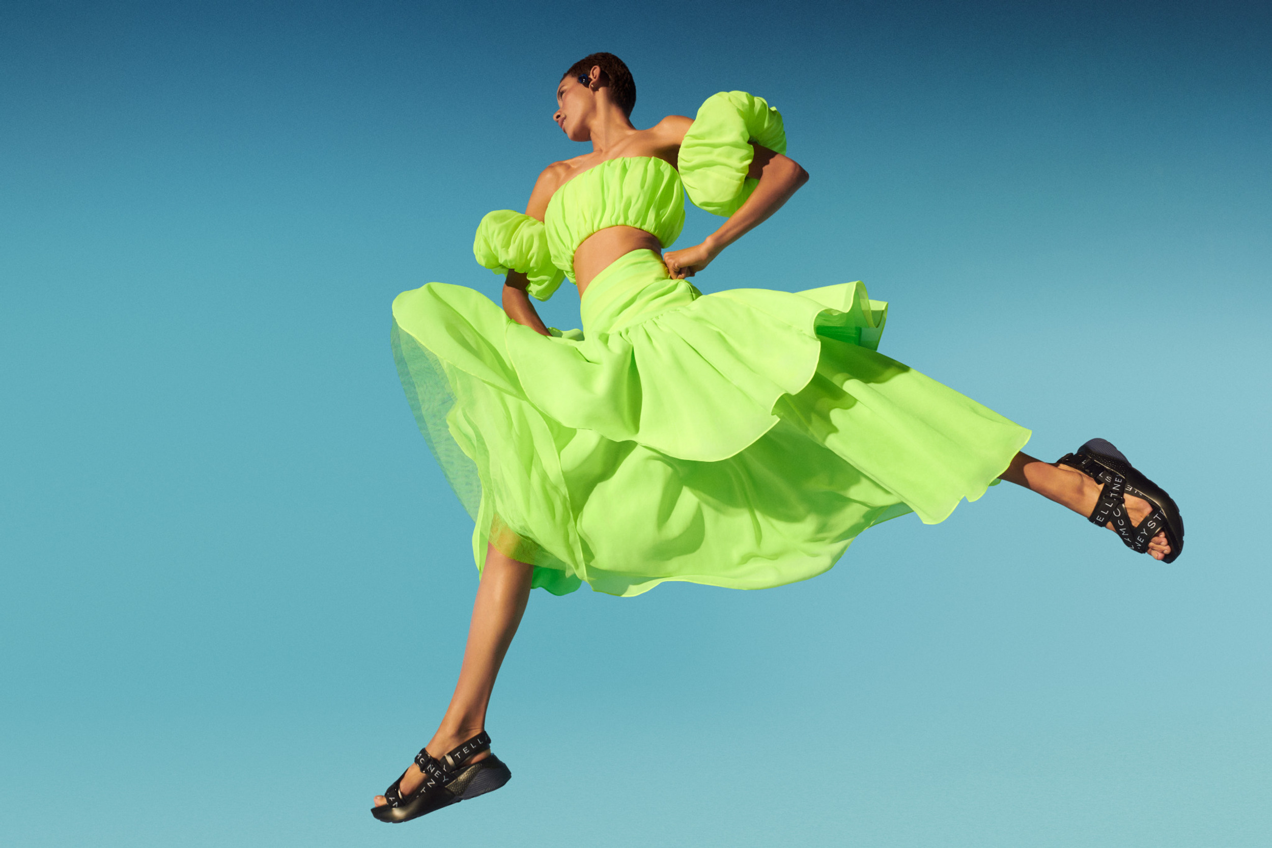 Fashion model in motion, wearing a neon green crop top and skirt with black sandals, set against a vibrant blue sky backdrop.