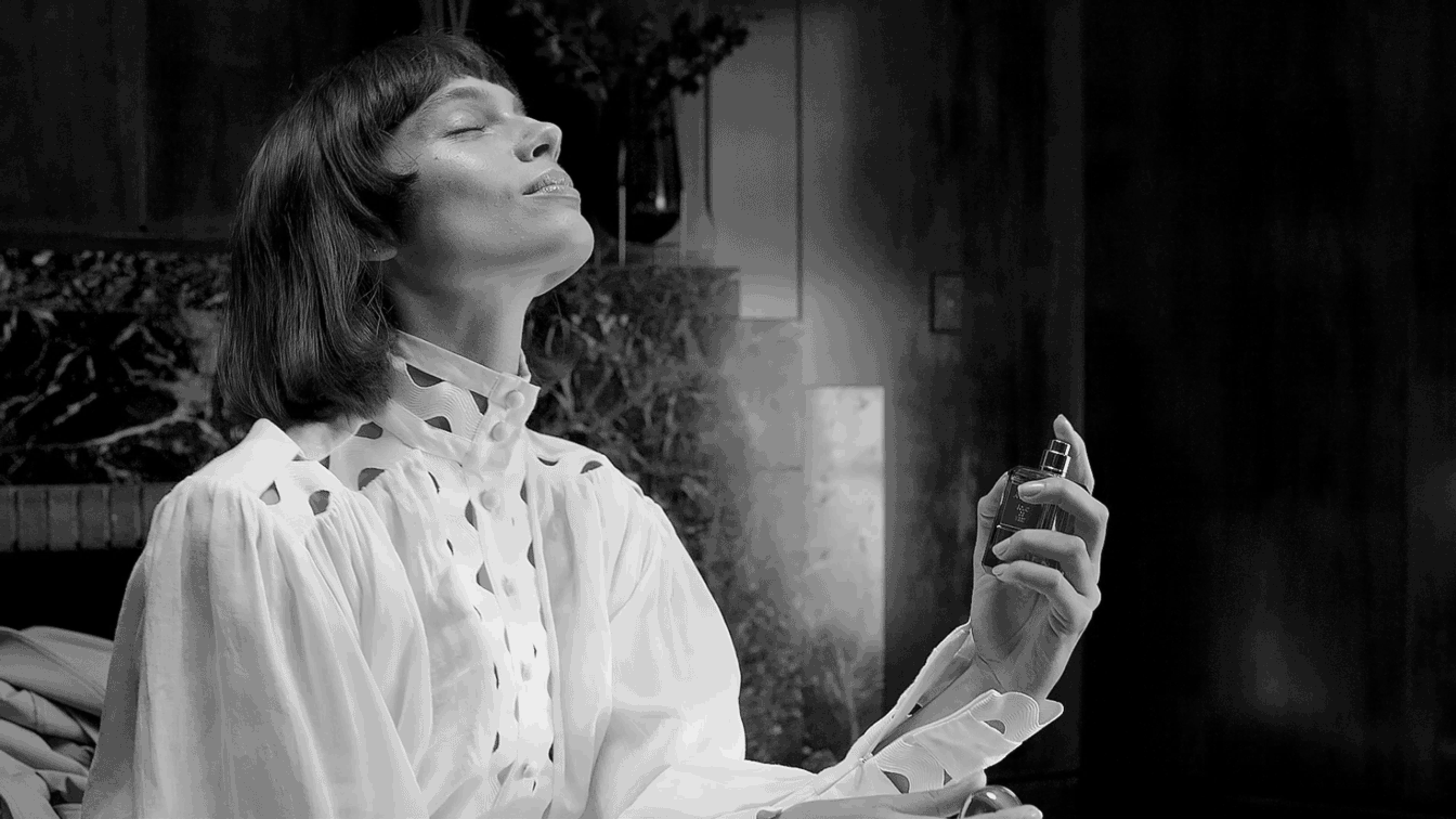 Monochrome portrait of a woman spraying perfume in an indoor environment with dark marble and a vase of plants in the background.