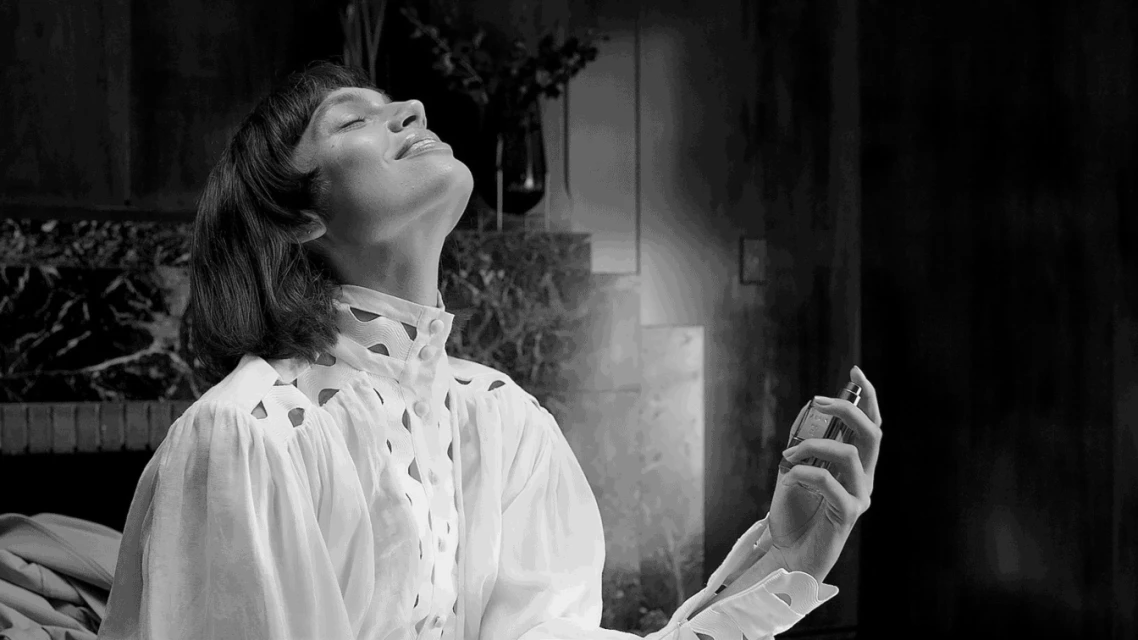 Monochrome portrait of a woman spraying perfume in an indoor environment with dark marble and a vase of plants in the background.