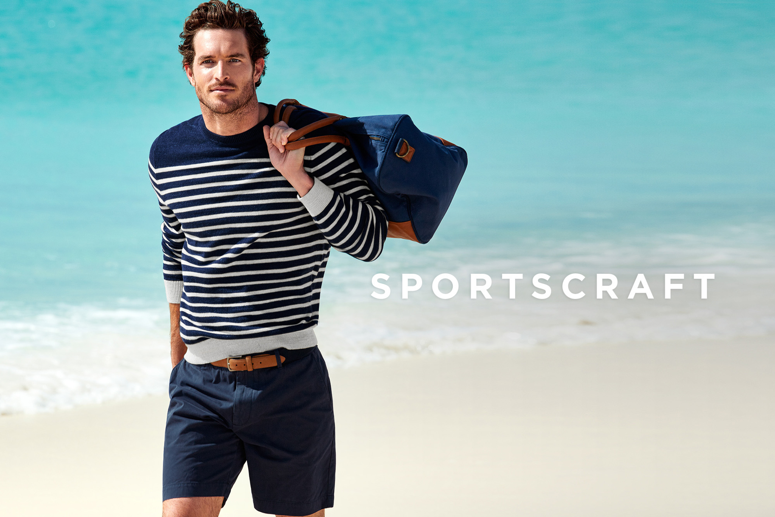 A light-skinned man in a striped sweater and shorts stands on a beach with a duffel bag against a blurred sea and sky background.