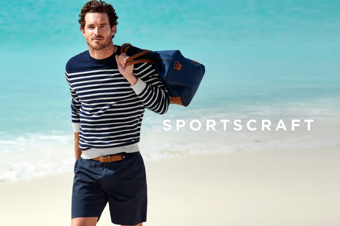 A light-skinned man in a striped sweater and shorts stands on a beach with a duffel bag against a blurred sea and sky background.