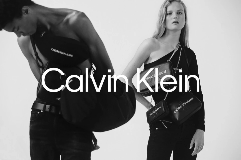 Monochrome studio shot of an adult male and female wearing Calvin Klein clothing and carrying bags. Brand logo prominently displayed.
