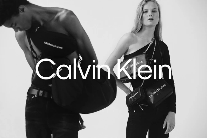 Monochrome studio shot of an adult male and female wearing Calvin Klein clothing and carrying bags. Brand logo prominently displayed.