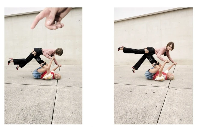 Two women perform an acrobatic pose outdoors with one lying on the ground and the other balancing on her legs, with a hand pointing down.