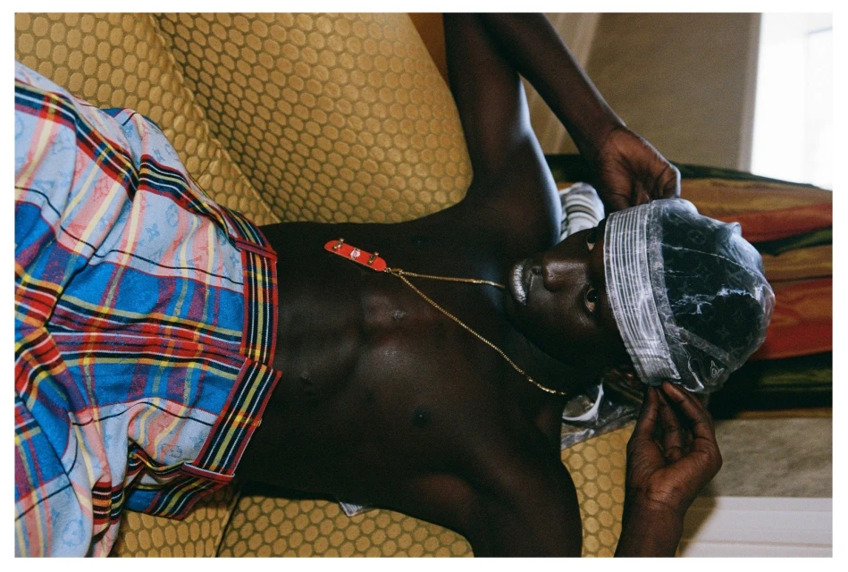 Shirtless Black man lies on a yellow patterned couch wearing a clear cap, plaid pants, and a gold chain with a skateboard charm.