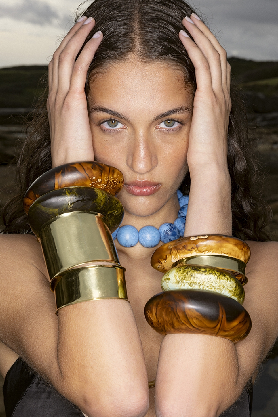 Close-up of a woman with curly hair holding her hands near her head, wearing a blue beaded necklace and large wooden and gold bracelets.