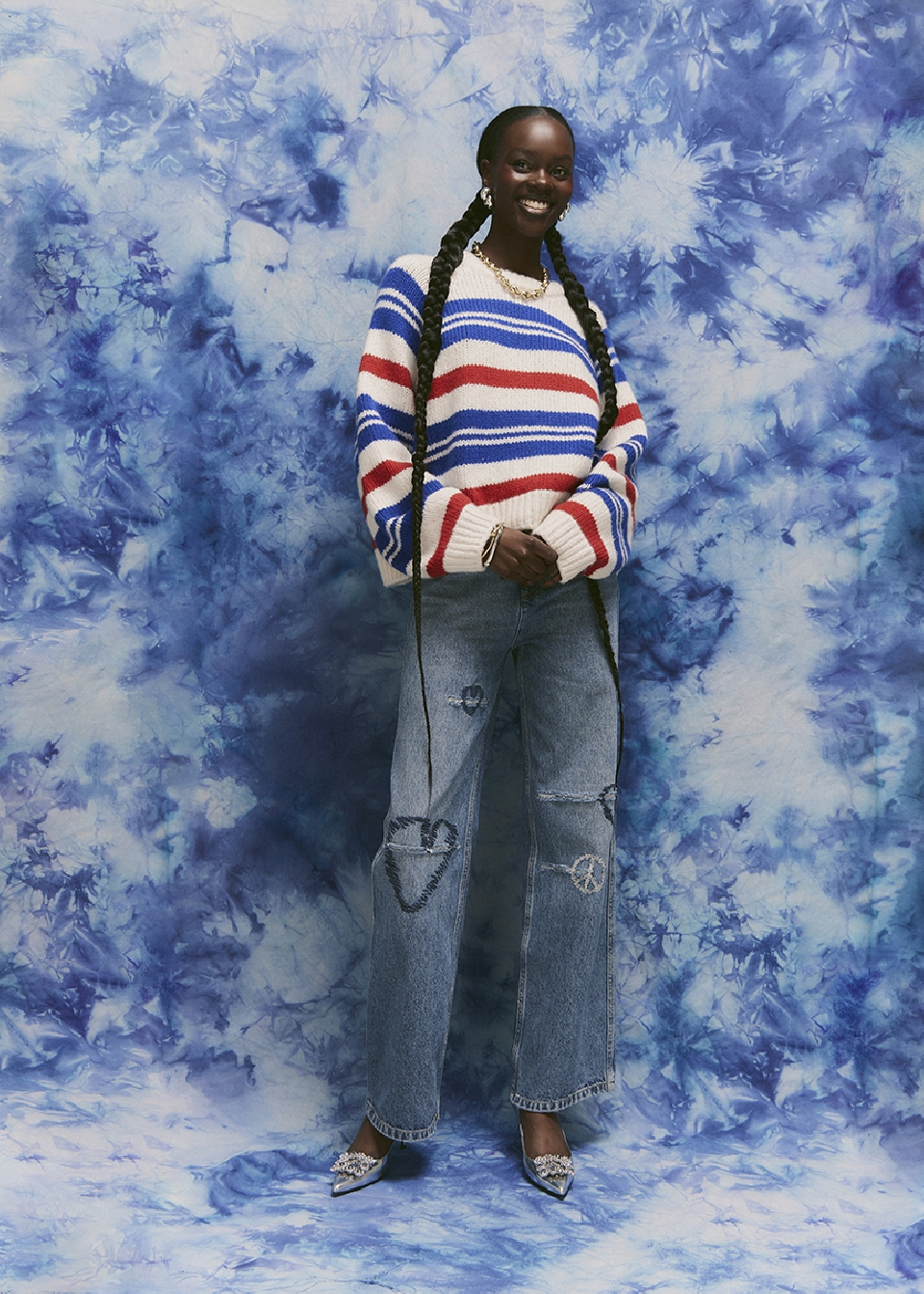 A smiling African woman in a striped sweater and jeans stands against a tie-dye backdrop. She wears a gold necklace and statement earrings.