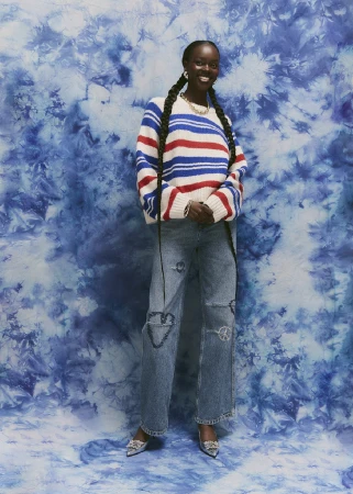 Black woman wearing a striped sweater and jeans with long braided pigtails standing against a blue tie-dye backdrop