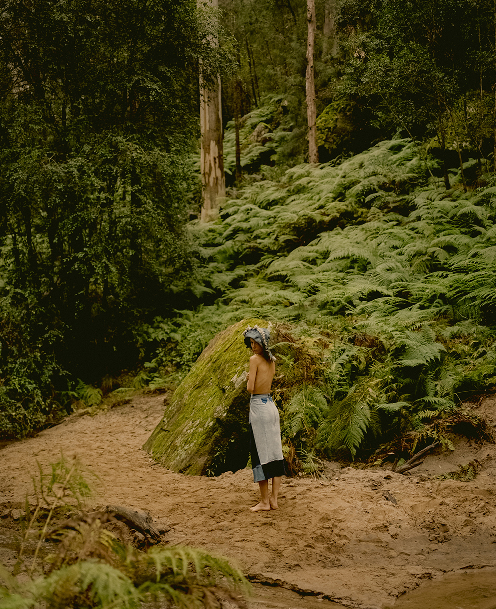 Woman in skirt standing on dirt path surrounded by green ferns and trees in dense forest, captured in a natural setting.