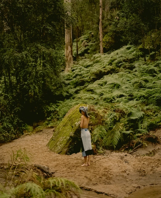 Woman in skirt standing on dirt path surrounded by green ferns and trees in dense forest, captured in a natural setting.