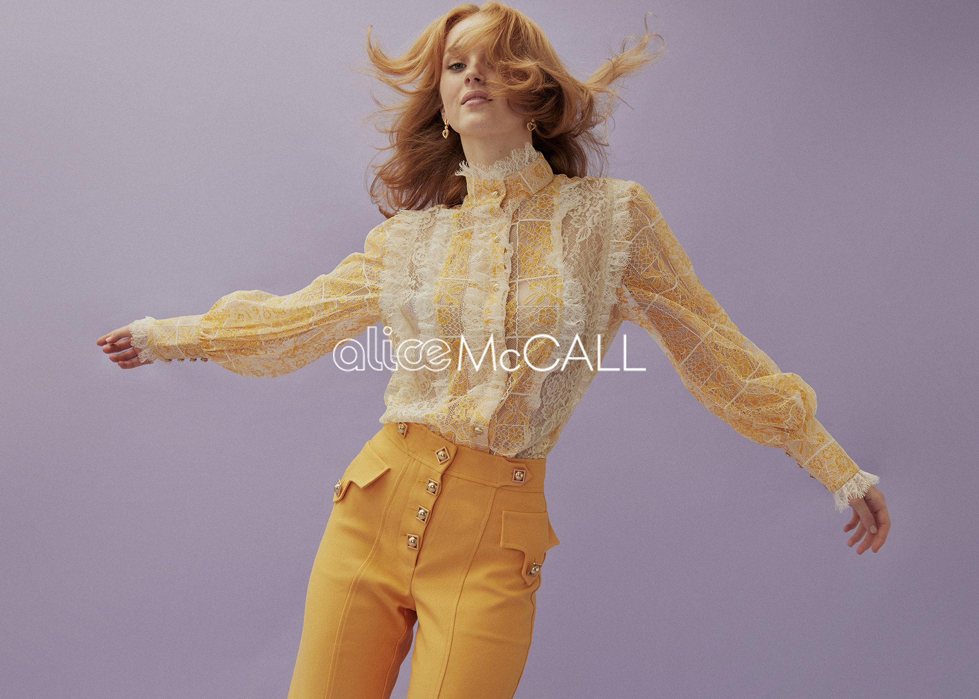 Fashion portrait of a red-haired female adult wearing a yellow and white lace top and high-waisted yellow pants against a lilac background.
