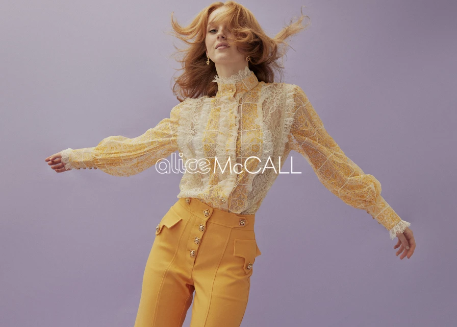 Fashion portrait of a red-haired female adult wearing a yellow and white lace top and high-waisted yellow pants against a lilac background.