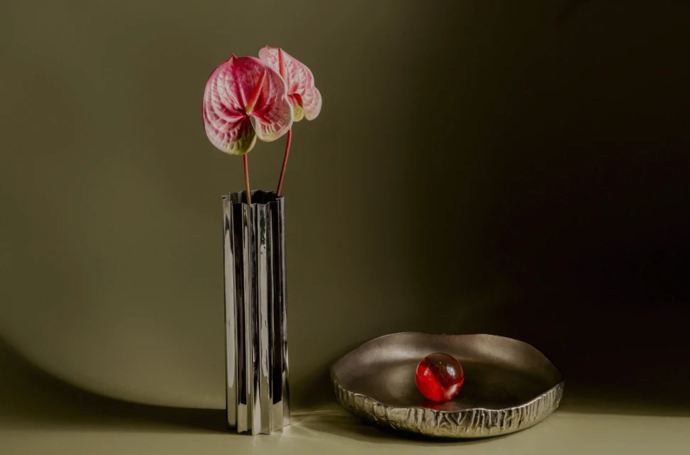 Still life photo of pink Anthurium flowers in a silver vase, a silver bowl with a red glass ball, against an olive green background.