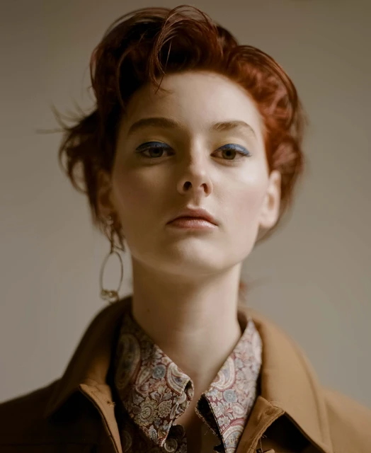 Close-up portrait of a young woman with auburn hair, blue eyeshadow, and a paisley-print shirt under a tan jacket.