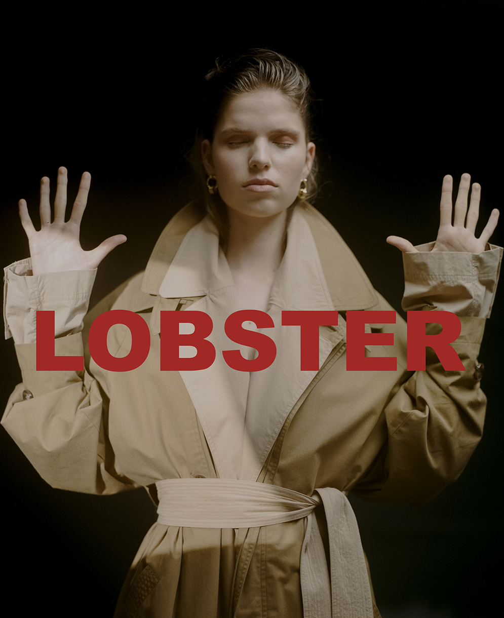 Woman with eyes closed wearing a tan trench coat with arms raised against a dark background, by photographer Natasha Killeen.