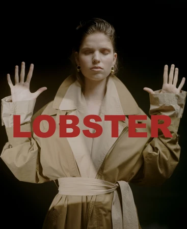 Woman with eyes closed wearing a tan trench coat with arms raised against a dark background, by photographer Natasha Killeen.