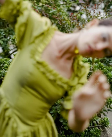 Woman in a light green dress strikes a dynamic pose amidst lush green foliage. Close-up shot focusing on her joyful expression.