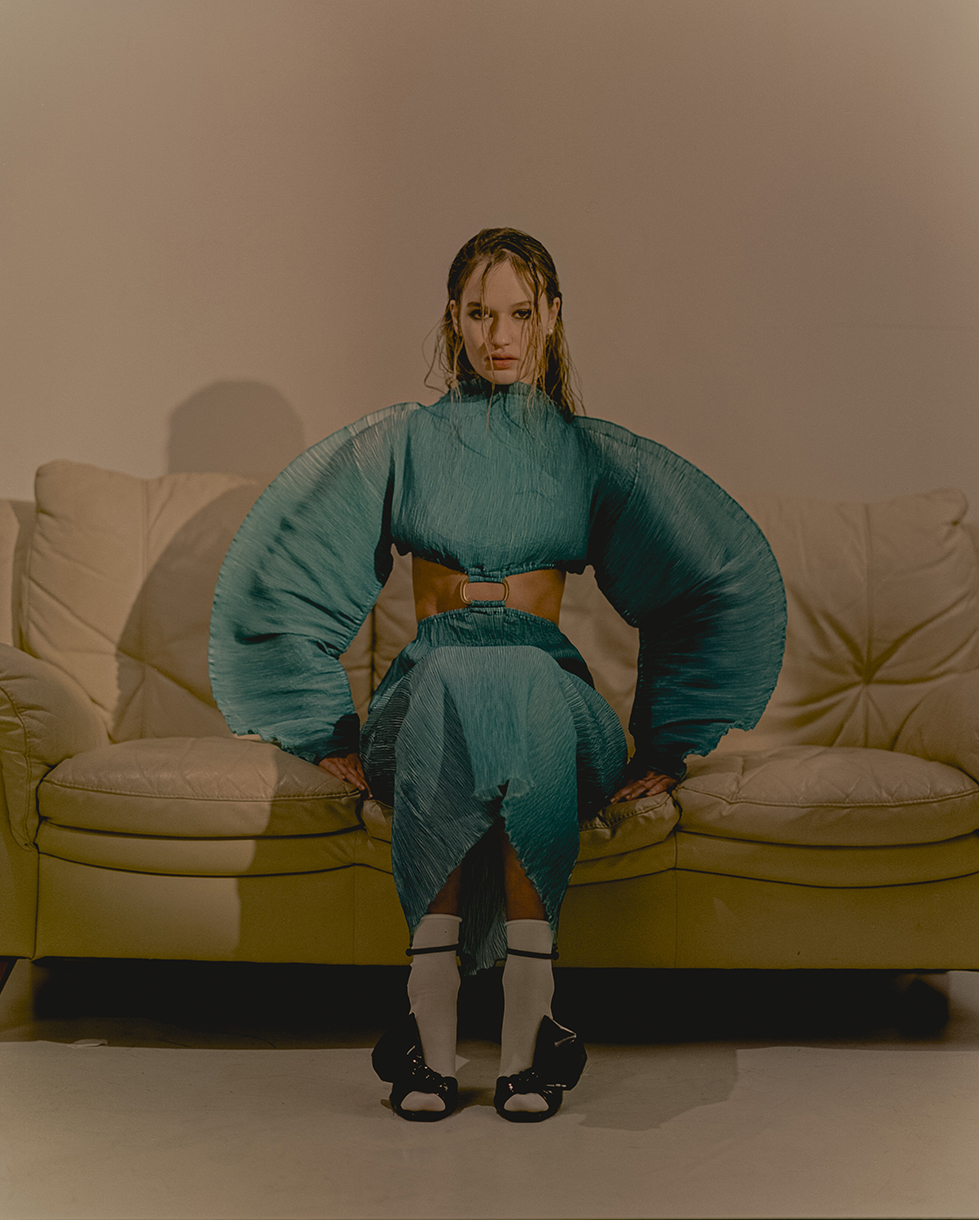 Woman with wet hair poses on a cream sofa in a matching teal pleated top and skirt ensemble, complete with white socks and black shoes.