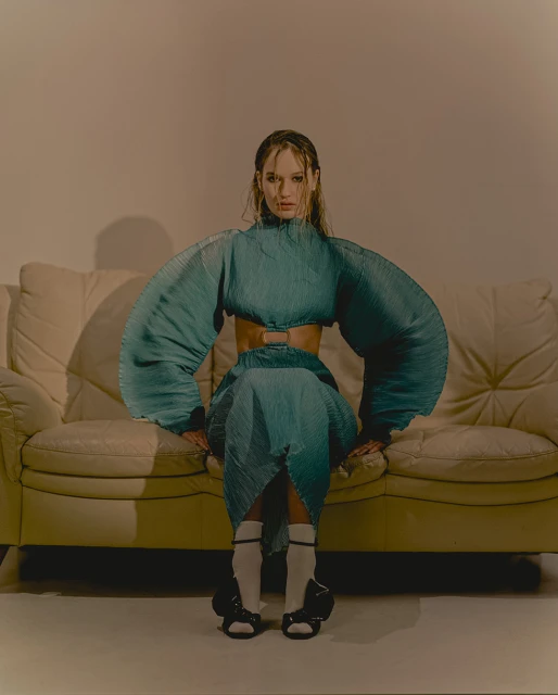 Woman with wet hair poses on a cream sofa in a matching teal pleated top and skirt ensemble, complete with white socks and black shoes.