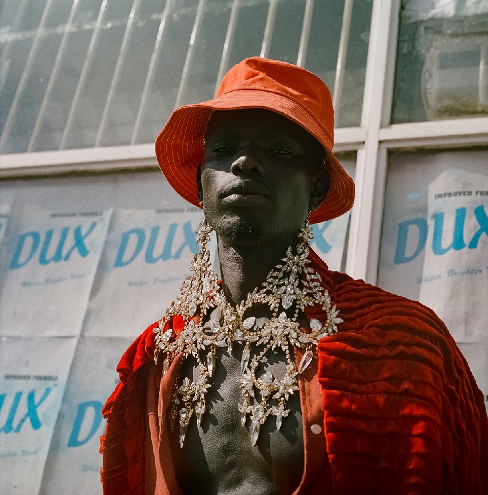 Portrait of a dark-skinned adult man wearing a coral bucket hat, large costume jewelry necklace, and a red velvet jacket, posing outdoors.