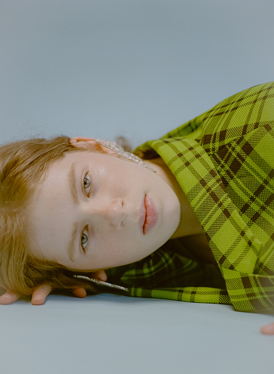 Close-up studio shot of a light-skinned young woman with light brown hair wearing a bright green plaid jacket and sparkling earrings.