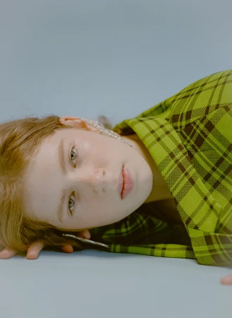 Close-up studio shot of a light-skinned young woman with light brown hair wearing a bright green plaid jacket and sparkling earrings.