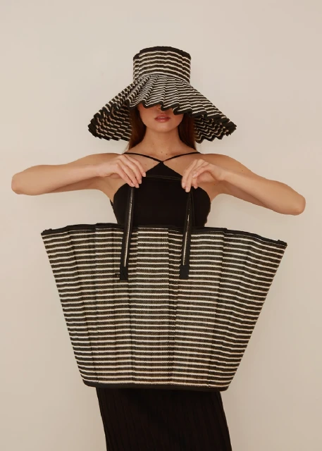 Woman wearing striped sun hat and holding a large striped tote bag. Studio shot with a neutral background. Fashion and style.