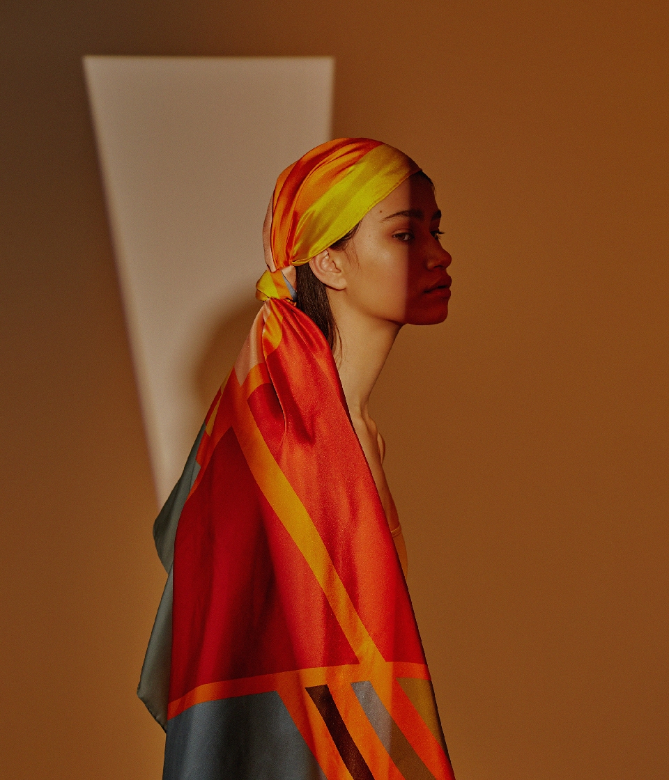 Profile shot of a young woman with a scarf on her head, against a warm orange background with a geometrical light accent. 
