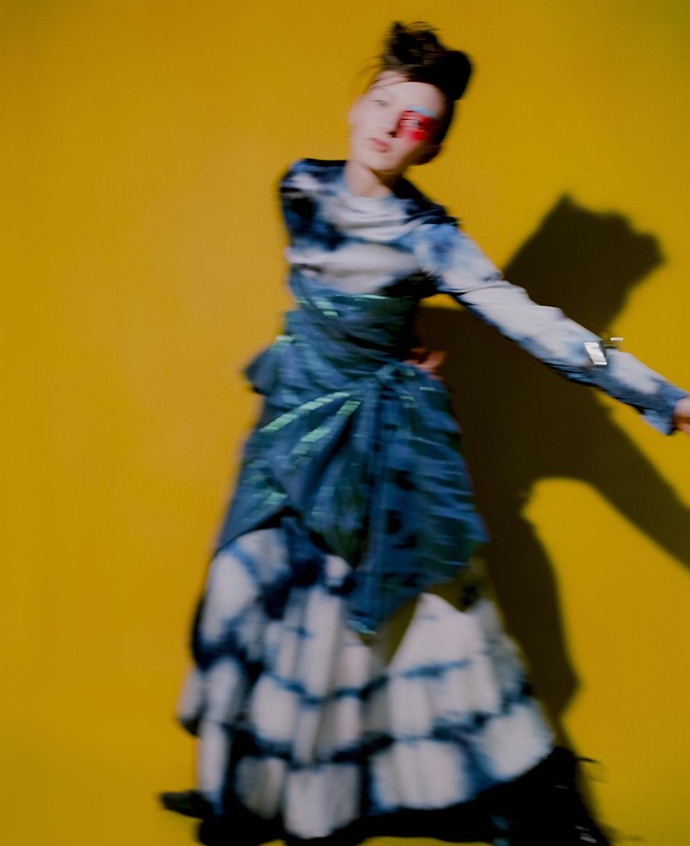 Woman in motion wearing blue-and-white dress against yellow backdrop, expressive makeup, dynamic fashion shot, studio lighting.