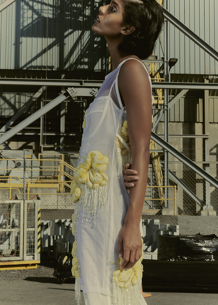 Adult woman poses outdoors in a sheer white dress with yellow flower accents, looking upward with an industrial background and natural light.