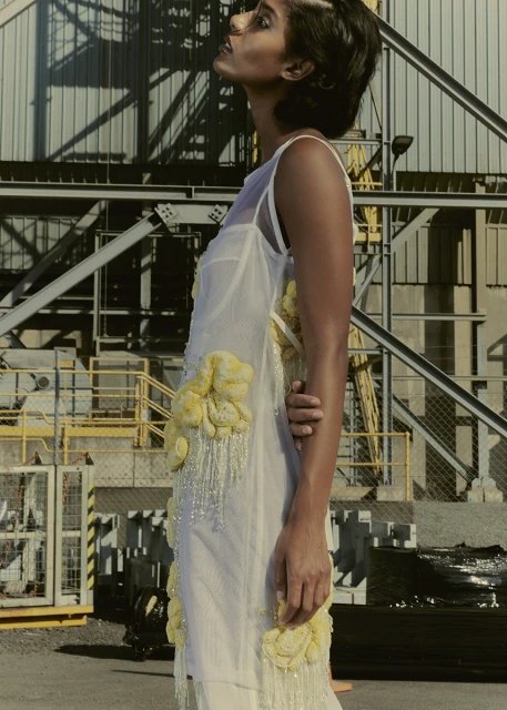 Adult woman poses outdoors in a sheer white dress with yellow flower accents, looking upward with an industrial background and natural light.