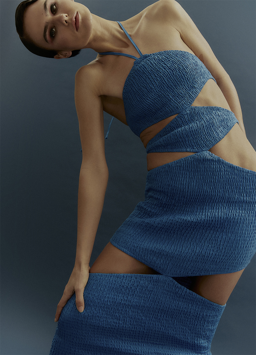 A woman poses in a studio wearing a textured, cut-out blue dress against a solid blue background, showcasing a modern and edgy fashion style.