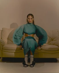 Woman in pleated teal long-sleeve top and matching skirt seated on a cream leather sofa, photographed by Natasha Killeen.