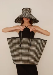 Model wearing a striped wide-brim hat and halter top holding a large striped tote bag against a plain white background.