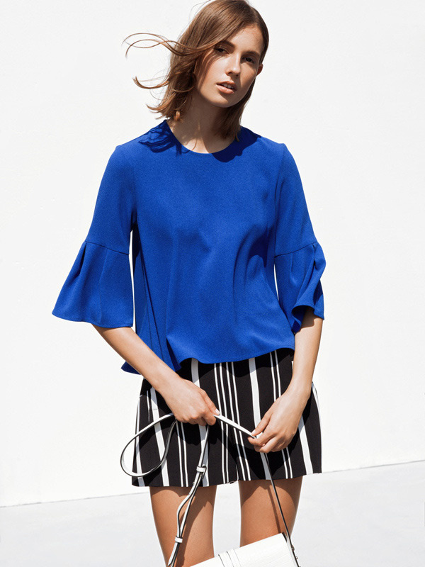 A woman models a vibrant blue blouse with bell sleeves and a black-and-white striped skirt against a clean white backdrop.