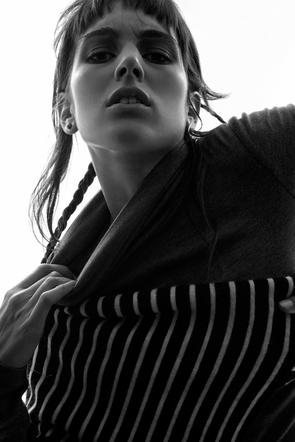 Black and white portrait from a low angle features a woman with braided hair pulling open a striped shirt, gazing intensely upward.