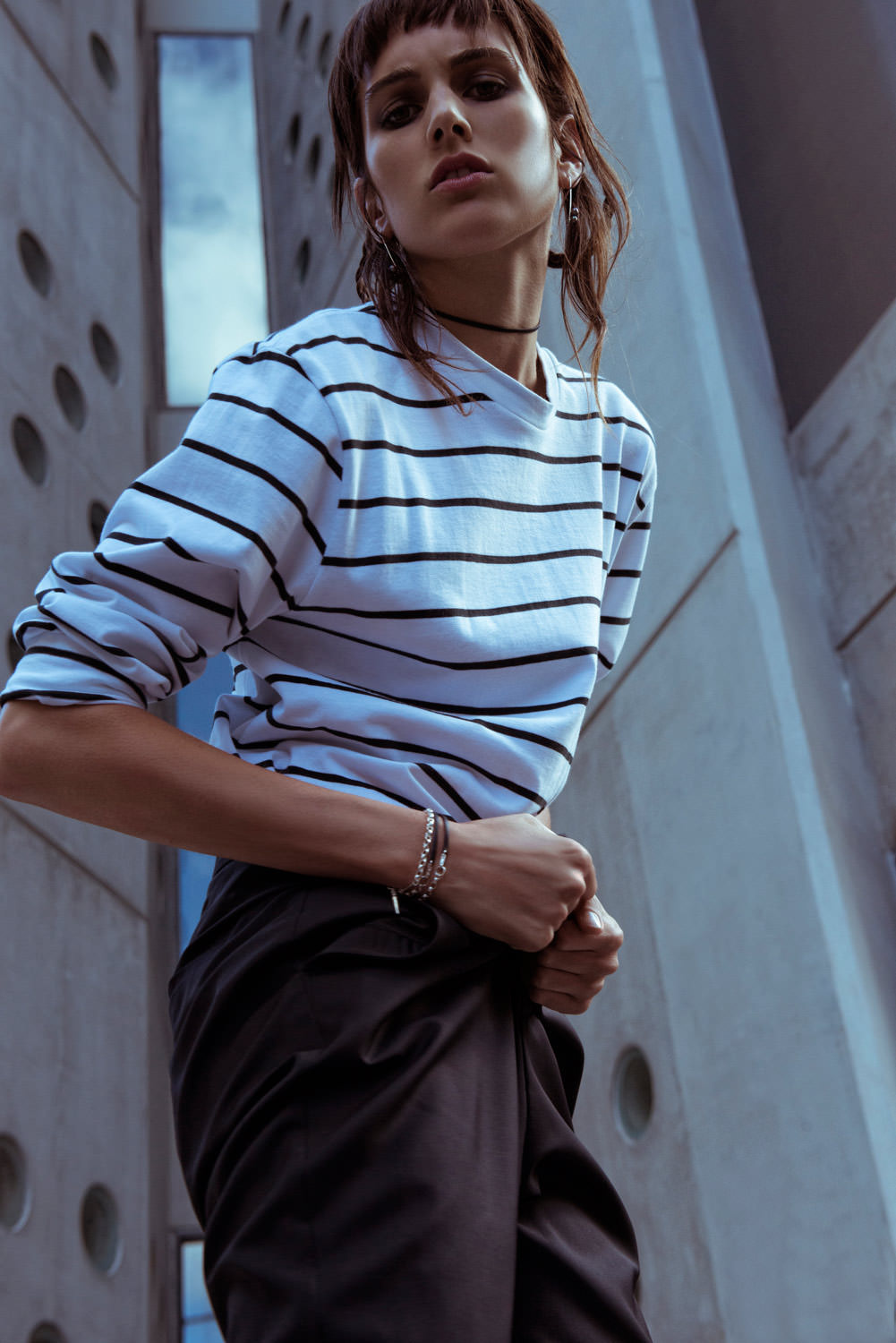 Fashion shot of a woman in a striped long-sleeve shirt and dark pants posing in front of a modern concrete building.