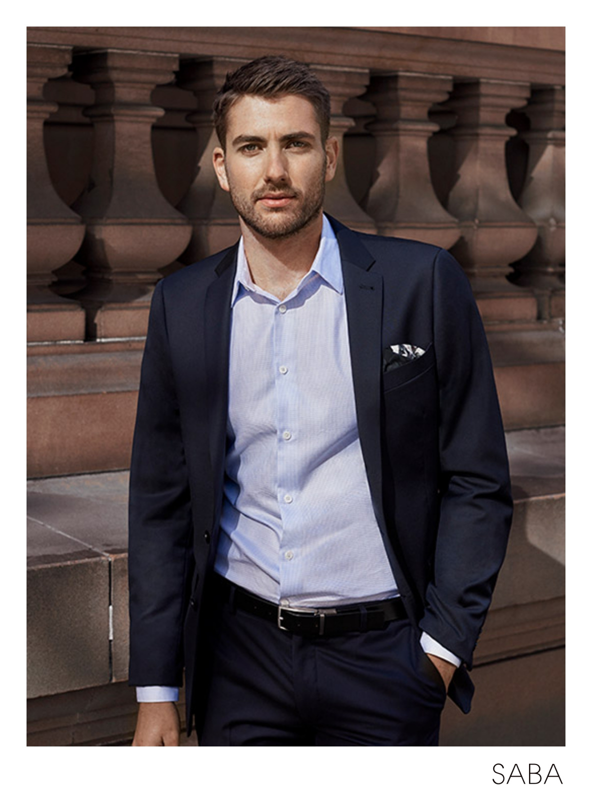 A handsome man in a navy suit and light blue shirt stands in front of a brown stone wall, looking confidently at the camera.