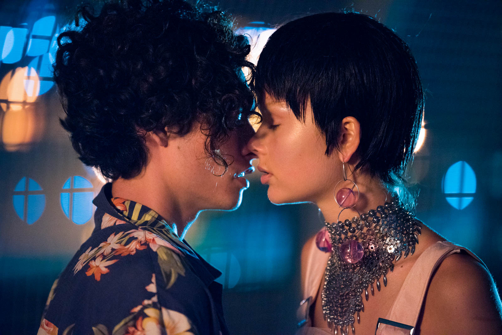 Close-up of a couple embracing, the male with curly hair and floral shirt, the female with a blunt haircut and statement jewelry against a blurred background.
