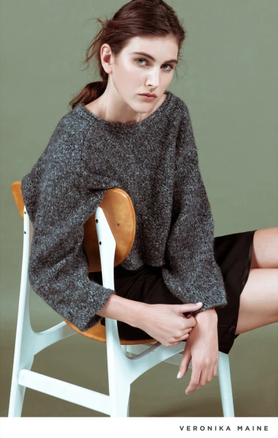 A woman with dark hair poses wearing a grey sweater and black skirt while sitting on a white and wooden chair in a studio.