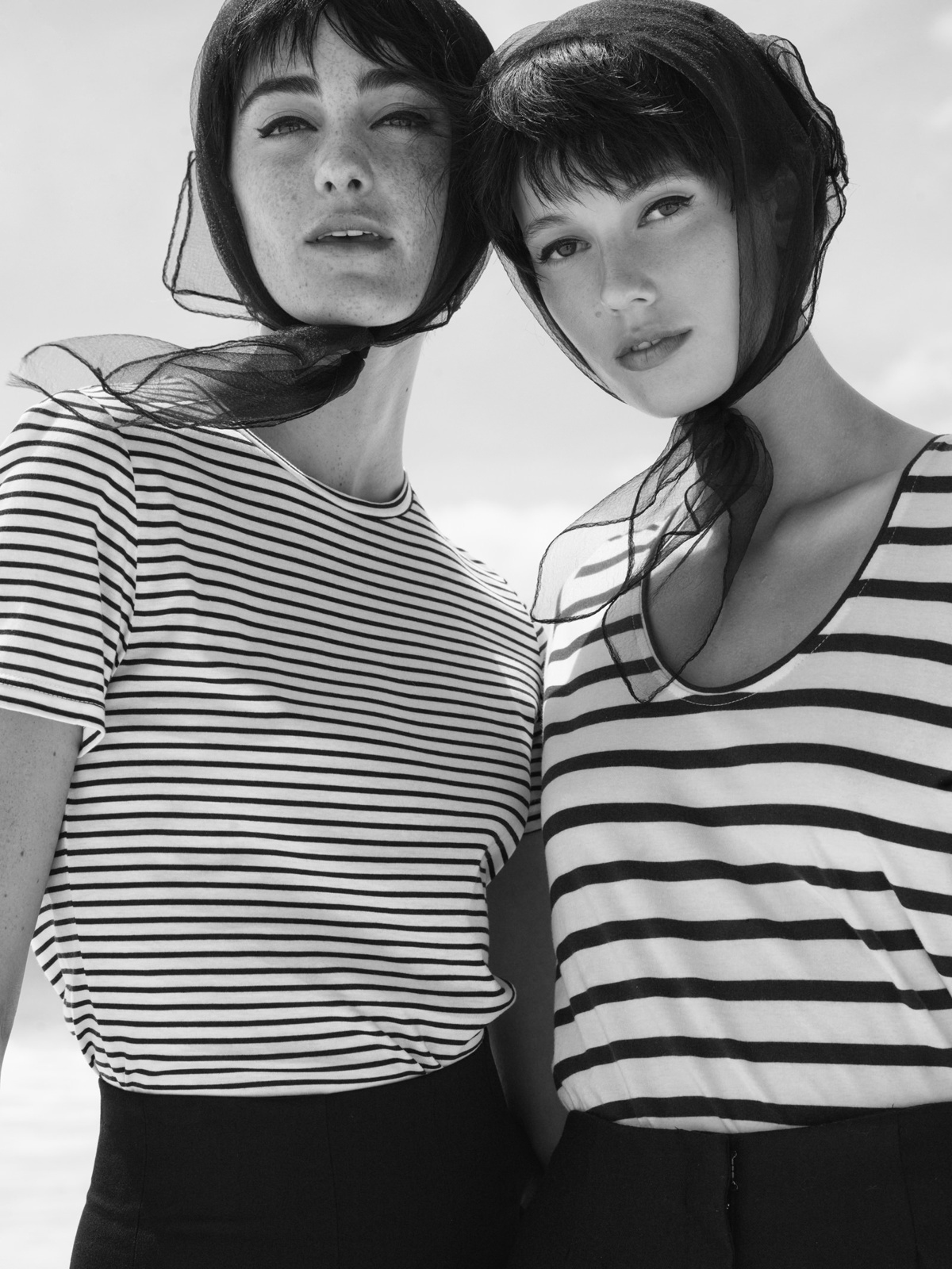 Black and white shot of two young women in striped tops and headscarves posing outdoors with a bright sky background, fashion.