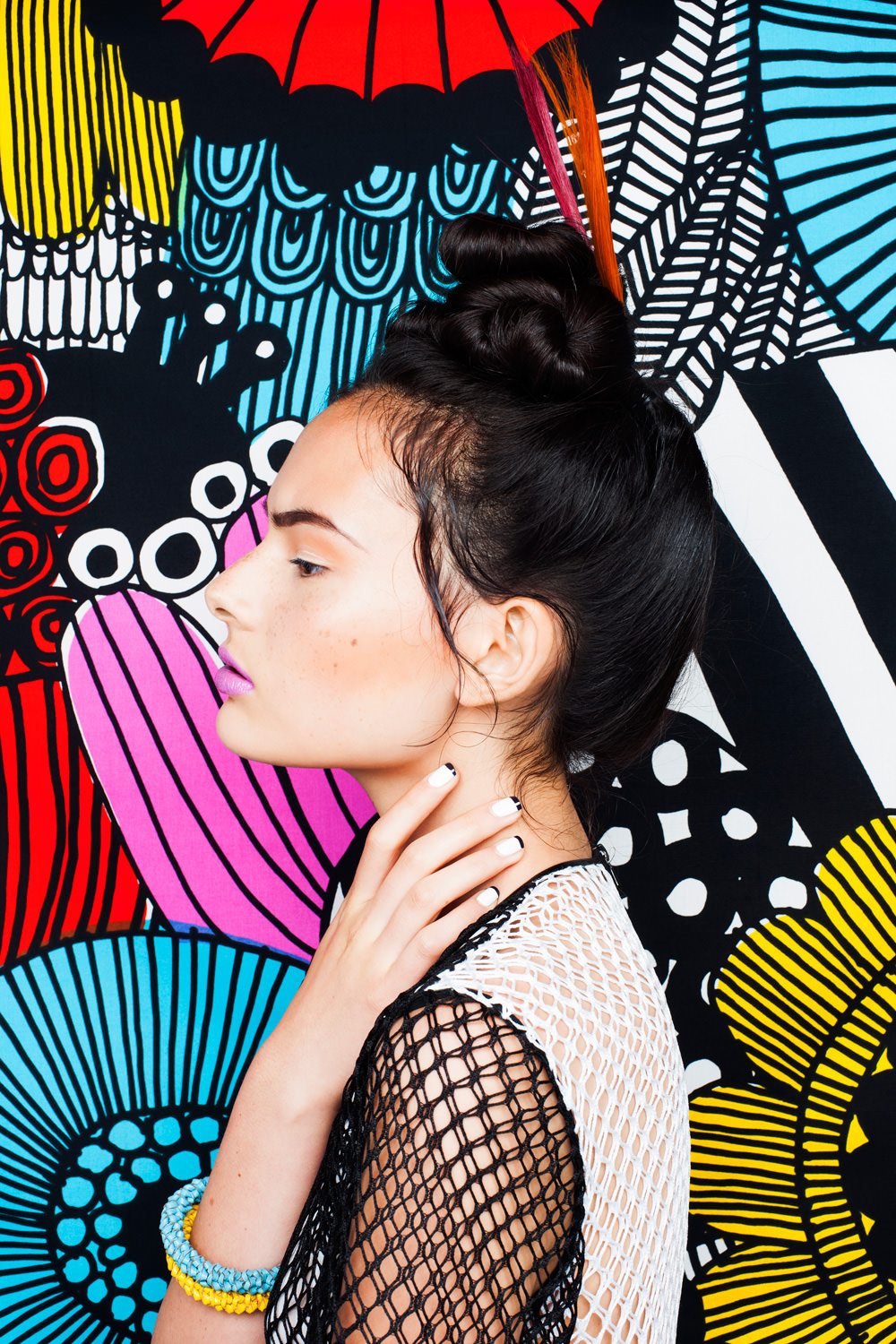 Profile shot of a model with updo hairstyle wearing a mesh top against a colorful abstract patterned backdrop.