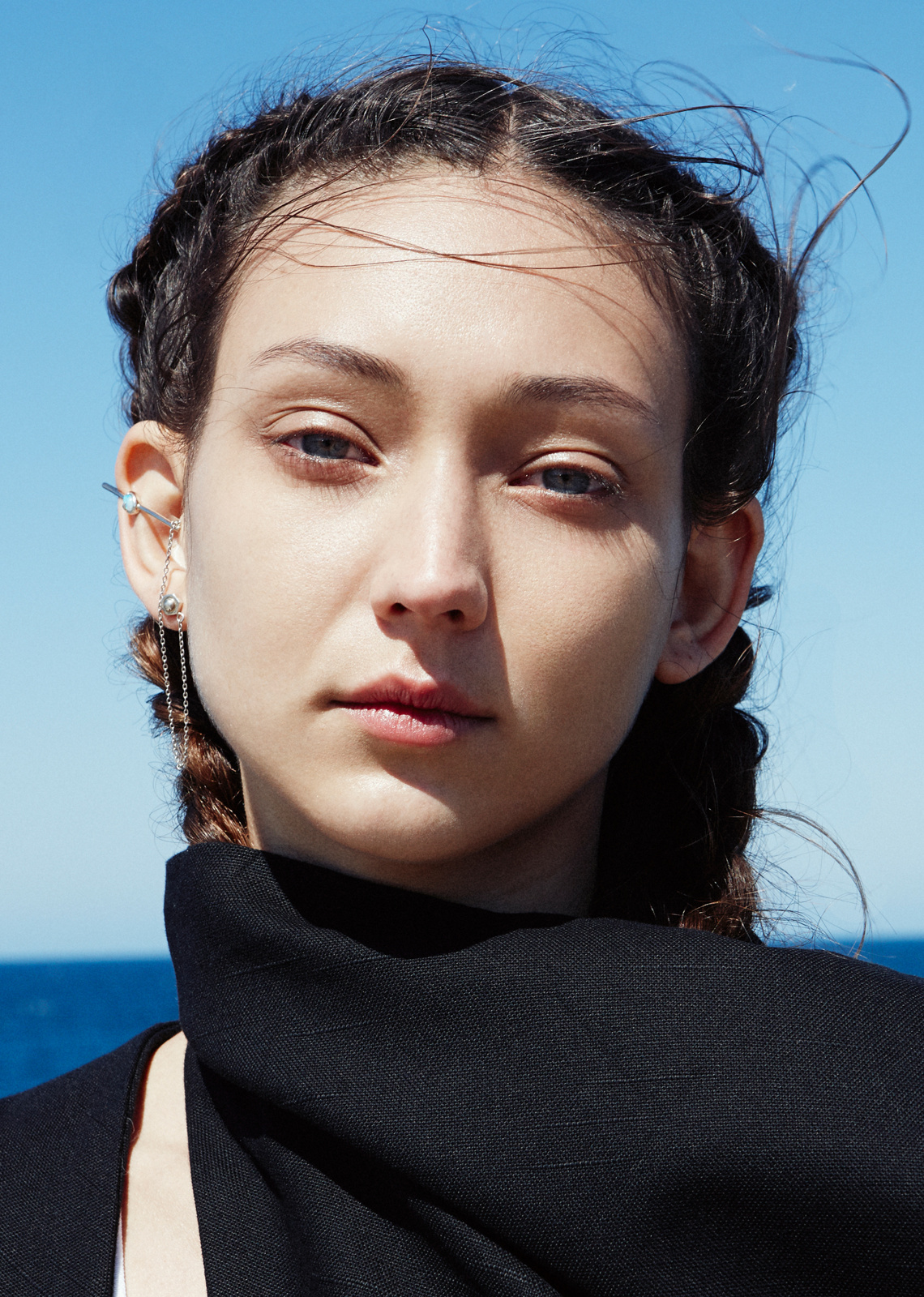 Close-up portrait of a young woman with braided hair wearing an ear cuff and black fabric against a sky and sea background.