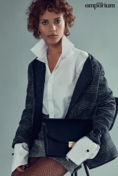 Model with short curly brown hair wearing a white collared shirt, plaid jacket, and holding a black handbag.