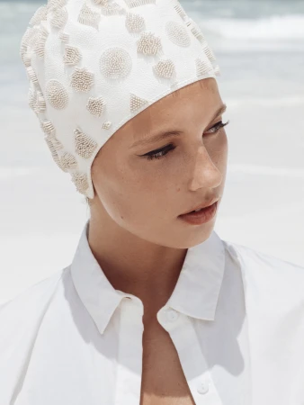 Model wearing a beaded white swim cap and white collared shirt against a bright sandy beach background